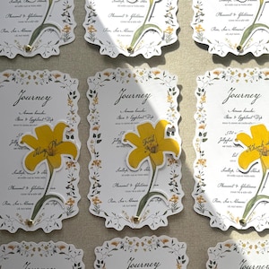 Op de afbeelding: Een verzameling rechthoekige menukaarten met de titel Journey, voorzien van een decoratieve bloemenrand en een gele lelie-illustratie. Elke kaart toont een gedrukte lijst met gerechten, waaronder Amuse bouche, Jellyfish, Scallop, Pheasant en Pear.