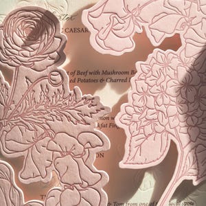 May include: Close-up of a light pink floral design on a cream-colored background. The design features embossed flowers and leaves, with the word "Menu" visible. The overall aesthetic is elegant and delicate.