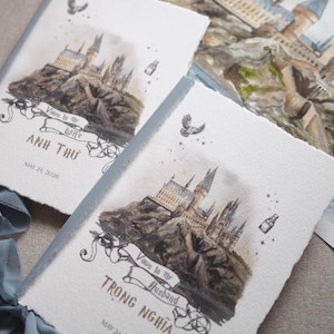 May include: Watercolor wedding vow books featuring a castle illustration. The books have a light blue ribbon and text that reads "Vows to my Wife" and "Vows to my Husband" with names and a date. The artwork is detailed and whimsical.