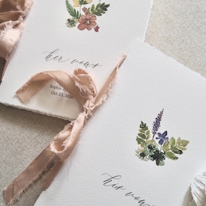 May include: Two white vow books with watercolor floral designs and calligraphy text. One book reads "her vows" and the other reads "his vows". Both books are tied with a peach ribbon. The names "Sophie & Jeffrey" and the date "Oct 23, 2026" are printed on the books.