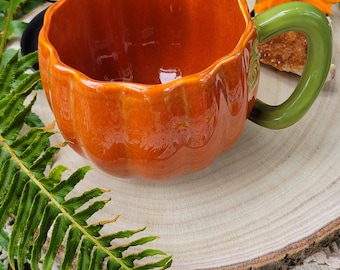 May include: Orange pumpkin-shaped ceramic mug with a green handle. The mug is sitting on a wooden slice and a white crocheted surface.