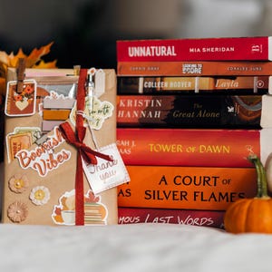 Puede incluir: Una pila de libros, incluyendo "Unnatural" y "A Court of Silver Flames", junto a un regalo con el texto "Bookish Vibes". Hojas de otoño y pequeñas calabazas completan la escena otoñal.
