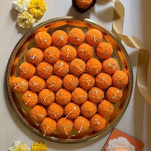 Pode incluir: Uma bandeja dourada adornada cheia de inúmeras guloseimas esféricas laranja, cada uma adornada com detalhes em folha de ouro. Os doces são dispostos em fileiras organizadas, sugerindo uma ocasião festiva ou comemorativa. Doces adicionais estão em xícaras de papel individuais. Flores amarelas e brancas e uma vela também estão presentes.
