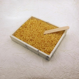 May include: A square, marble-look tray filled with small, yellow wax beads. A wooden applicator stick rests inside the tray, partially submerged in the wax. The tray sits on a textured, white surface.