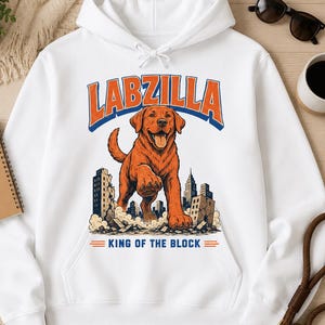 May include: A white hooded sweatshirt featuring a graphic of a large brown dog standing over a city skyline. The text above the dog reads LABZILLA in orange and blue, and the text below reads KING OF THE BLOCK in blue with orange lines.