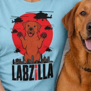 May include: Pale blue t-shirt featuring a dog graphic with the text "LABZILLA". The design includes a red sun and helicopters. A brown dog is next to the person wearing the shirt.