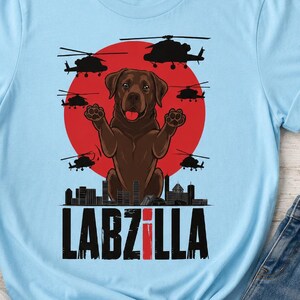 May include: A light blue t-shirt featuring a graphic of a brown Labrador dog with its paws raised, set against a large red circle. The design includes black silhouettes of helicopters, a city skyline, and the text LABZILLA in bold black letters.