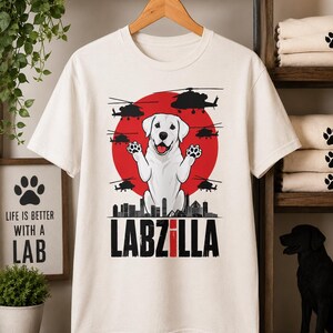 May include: A cream-coloured t-shirt featuring a graphic of a white Labrador retriever with raised paws in front of a large red circle. The design includes black silhouettes of helicopters, a city skyline, and the text LABZILLA in bold black letters.