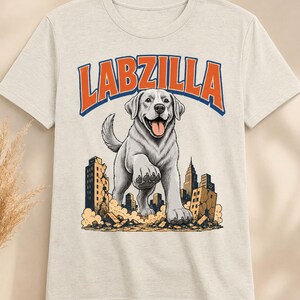 May include: A light beige short-sleeve t-shirt featuring a graphic print of a large Labrador dog walking through a city with crumbling buildings. The text LABZILLA is printed in bold orange letters with a blue outline above the dog illustration.