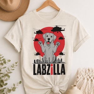 May include: A cream-coloured t-shirt featuring a graphic of a Labrador dog with raised paws in front of a large red circle. The design includes black silhouettes of helicopters, a city skyline, and the text LABZILLA printed in bold black letters.