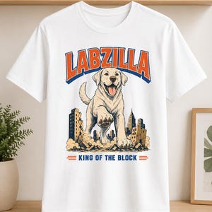 May include: A white short-sleeve t-shirt featuring a graphic of a large Labrador retriever standing over a city skyline. The text LABZILLA appears in orange and blue above the dog, with the text KING OF THE BLOCK below the city scene.
