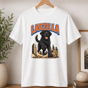 May include: A white short-sleeve t-shirt featuring a graphic of a large black Labrador dog standing over a crumbling city. Above the dog, the text LABZILLA is printed in orange with a blue outline. The dog has its tongue out and paws forward.