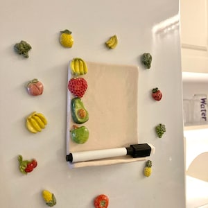 Puede incluir: Una puerta de nevera blanca adornada con imanes coloridos de frutas y verduras. Una pizarra rectangular beige con un rotulador blanco está adjunta. Imanes con forma de fresa, aguacate, manzana y otros productos rodean la pizarra.