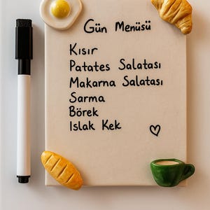 Puede incluir: Una pizarra de menú beige con el texto "Gün Menüsü" y una lista de platos turcos. Los imanes decorativos de alimentos incluyen un huevo frito, un cruasán, pan y una taza de café verde. Un rotulador blanco y negro está a la izquierda.
