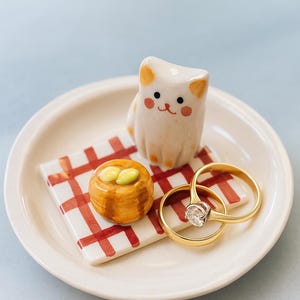 Op de afbeelding: Een keramische ringschaal met een wit kattenbeeldje, een klein gebakje en twee gouden ringen. De schaal heeft een rood-wit geblokt patroon. De ringen hebben een heldere edelsteen.