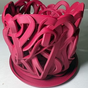 May include: A magenta-colored, 3D-printed planter with an intricate, openwork design. The planter has a cylindrical shape with a matching saucer base. The sides feature a repeating pattern of curved lines and shapes, creating a decorative and modern aesthetic.