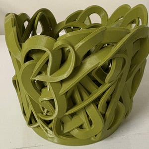 May include: A 3D-printed, olive-green decorative bowl with an intricate, interwoven design. The openwork pattern creates a visually interesting texture, suitable for holding small items or as a standalone art piece. The bowl's form is round with a slightly flared top.