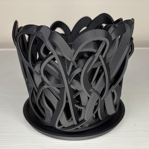 May include: A black 3D-printed planter with an intricate, interwoven design. The planter sits on a matching saucer, creating a cohesive look. The openwork design allows for air circulation, making it suitable for various plants. The planter is approximately 5 inches in diameter.