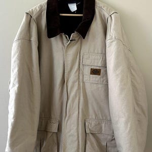 May include: A beige canvas work jacket featuring a dark brown corduroy collar, a front zipper closure, and two large lower flap pockets. A smaller chest pocket displays a rectangular Dickies brand patch. The jacket is hung on a wooden hanger.