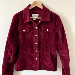 May include: A deep burgundy corduroy jacket featuring a classic collar, long sleeves, and a front button closure. The garment includes two chest pockets with buttoned flaps and a label at the inner neckline that reads A.M.I. with star symbols.