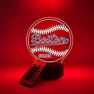 Pode incluir: Uma lâmpada LED circular com um gráfico de basebol, o texto Boston e o nome SHANE impresso no centro. A lâmpada está iluminada em vermelho e inclui um pequeno controlo remoto retangular com vários botões para definições de luz.