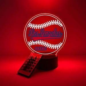 Pode incluir: Uma lâmpada LED circular de acrílico com um gráfico de uma bola de beisebol branca com costuras vermelhas. O texto Los Angeles está escrito em azul com um sublinhado azul, e o nome SHANE está impresso em letras azuis abaixo da bola.