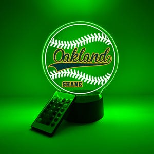 Pode incluir: Uma lâmpada LED circular de acrílico com um gráfico de basebol com o texto Oakland e o nome SHANE impressos no centro. A lâmpada emite uma luz verde brilhante e inclui um pequeno controlo remoto preto para ajustar as definições de luz.