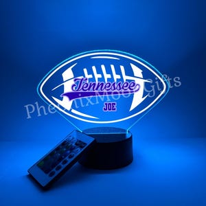 May include: A blue LED lamp shaped like a football, featuring the word "Tennessee" and the name "JOE" in white and purple. It comes with a remote control. The lamp is on a black base.