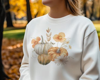 Könnte beinhalten: Cremefarbenes Sweatshirt mit einem Aquarelldesign aus Kürbissen, Blumen und Blättern in Orange-, Weiß- und Gelbtönen. Das Design umfasst einen Schmetterling und zarte florale Akzente, perfekt für den Herbst.