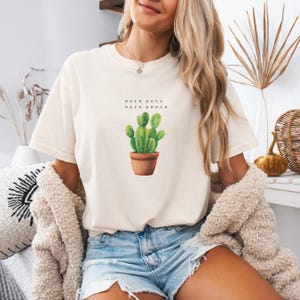 Puede incluir: Camiseta color crema con una ilustración de acuarela de un cactus verde en una maceta marrón. El texto "more pots more peace" está impreso encima del cactus. La camiseta se combina con pantalones cortos de mezclilla y una rebeca beige.