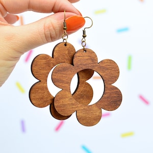 Puede incluir: Pendientes de madera marrón con forma de flor y diseño retro. Los pendientes tienen forma de flor recortada. Los sostiene una mano con esmalte de uñas naranja sobre un fondo blanco con confeti de colores.