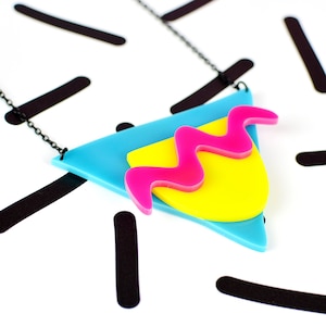 May include: A turquoise, yellow and pink geometric necklace with a black chain. The pendant is a triangle shape with a wavy pink design on top of a yellow rectangle.