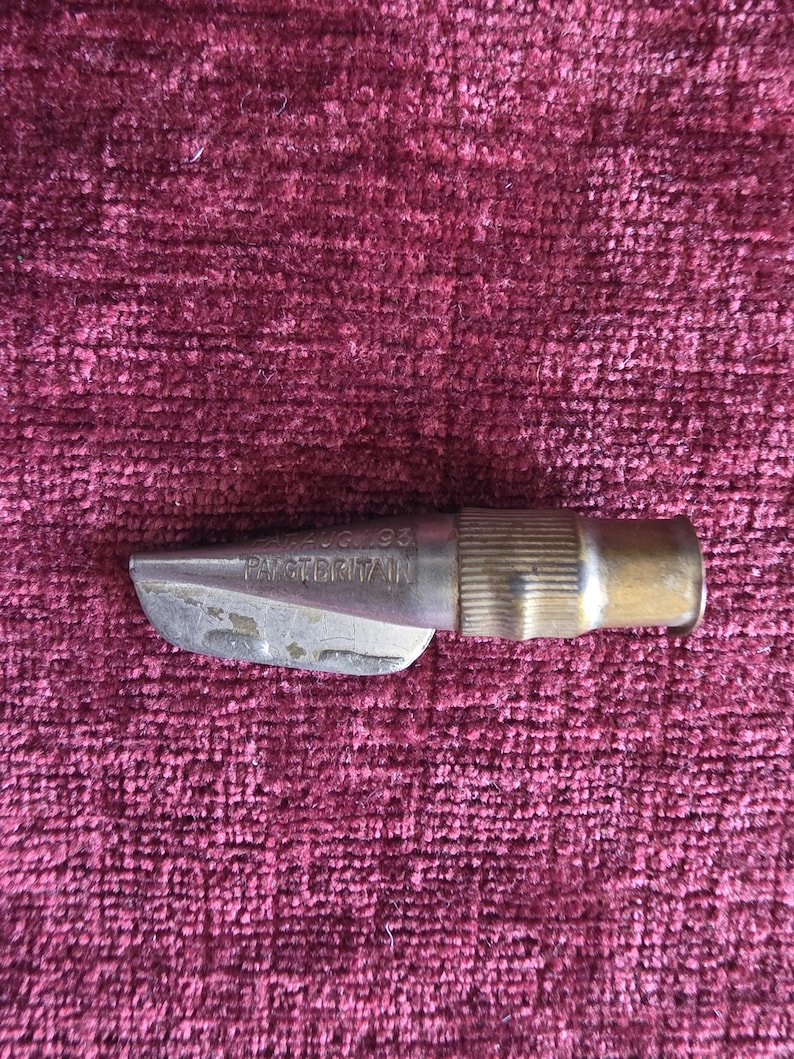 K&ouml;nnte beinhalten: Ein metallisches Werkzeug mit einer silberfarbenen Spitze und einem gerippten, messingfarbenen zylindrischen Sockel. Der silberne Teil tr&auml;gt die eingepr&auml;gte Aufschrift PAT AUG 93 und PAT GT BRITAIN. Das Objekt hat eine konische Form.