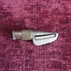 K&ouml;nnte beinhalten: Ein silberfarbenes Vintage-Metallwerkzeug mit einem zylindrischen, gerippten Griff und einem konisch zulaufenden Klingengeh&auml;use. An der Seite des Metallk&ouml;rpers befindet sich der gepr&auml;gte Schriftzug THE BELLE. Das Objekt weist Gebrauchsspuren auf.