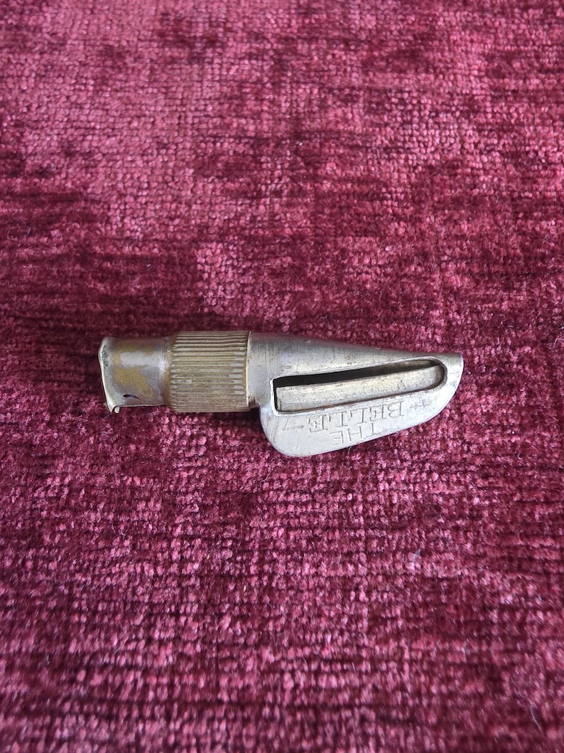 K&ouml;nnte beinhalten: Ein silberfarbenes Vintage-Metallwerkzeug mit einem zylindrischen, gerippten Griff und einem konisch zulaufenden Klingengeh&auml;use. An der Seite des Metallk&ouml;rpers befindet sich der gepr&auml;gte Schriftzug THE BELLE. Das Objekt weist Gebrauchsspuren auf.