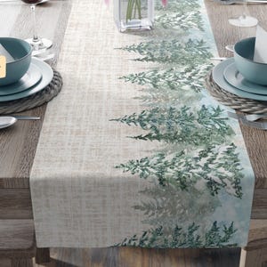 May include: A polyester table runner with a winter forest design. The runner features a light beige background with a pattern of green and white evergreen trees. The table is set with teal bowls, plates, and silverware.