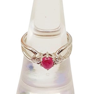 Op de afbeelding: Een zilveren ring met een centrale ovaalvormige, dieprode edelsteen, geflankeerd door kleinere, heldere stenen. De band van de ring heeft een gespleten ontwerp, waardoor hij een elegante en delicate uitstraling heeft. De ring wordt getoond op een heldere, parelmoerachtige standaard.