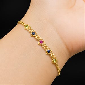 Può includere: Un bracciale dorato con maglie a forma di cuore, ognuna con una gemma colorata. Le gemme sono blu, rosa e verdi. Il bracciale è indossato su un polso, mostrando il suo design delicato e i colori vivaci.