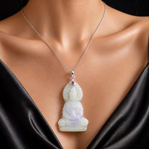 Può includere: Una collana d'argento con un ciondolo in giada a forma di Buddha seduto. Il ciondolo è verde chiaro e lavanda, con una piccola gemma rossa in cima. La collana è esposta su uno sfondo nero.