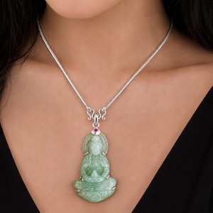 May include: A light green carved jade pendant in the shape of a seated figure, suspended from a silver snake chain. The pendant features a small, round, deep pink gemstone set at the top bail, connecting the jade carving to the metallic chain.