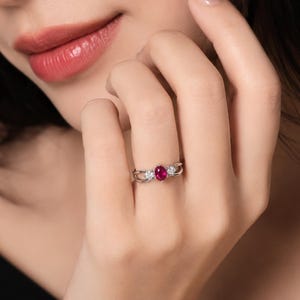 Könnte beinhalten: Ein silberfarbener Ring mit einem zentralen, oval geschliffenen, leuchtend roten Edelstein, der von zwei kleineren, rund geschliffenen, klaren Steinen flankiert wird. Die Ringschiene ist gespalten und umschließt die Steine elegant.