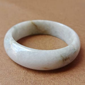 May include: A smooth, polished ring with a rounded profile, crafted from a pale, translucent stone featuring subtle, cloudy white tones and faint, natural brown inclusions that create a unique, organic pattern throughout the band's surface.