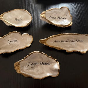 Puede incluir: Cinco tarjetas de lugar con forma de concha de ostra con bordes dorados. Las tarjetas son blancas con escritura negra que dice "Bride", "Groom", "Nina Banks", "Annie Banks-MacKenzie" y "George Banks".