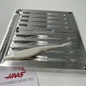 May include: A silver metal mould with multiple cavities, holding a white, fish-shaped lure. The mould has a rectangular shape with a channel on the left side. A paper with the text "HAAS HIGH-PERFORMANCE MACHINE TOOLS" is at the bottom.