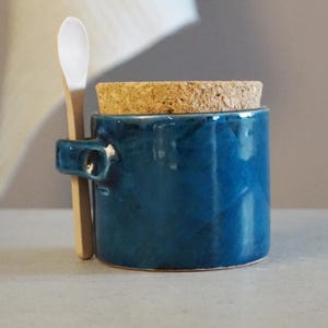Peut inclure: Petit pot rond en céramique bleue avec un couvercle en liège et une petite poignée. Une petite cuillère en bois est posée contre le côté du pot. Le pot mesure environ 7,6 cm de haut.
