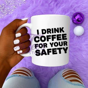 May include: White ceramic mug with the text "I DRINK COFFEE FOR YOUR SAFETY" in black. The mug is held against a purple fuzzy background. A purple disco ball and a white sphere are also visible.