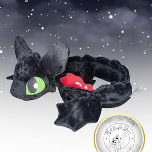 Può includere: Un peluche di drago nero con occhi verdi e interno rosso. Il drago ha un corpo lungo, ali e una testa dettagliata. Lo sfondo presenta un cielo stellato.
