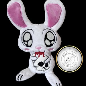 May include: A white plush rabbit toy with large eyes, pink inner ears, and pink cheeks. The rabbit has red embroidery around its mouth resembling blood and is holding a white skull. A circular logo with the text Mad Knight Dream Designs is visible.