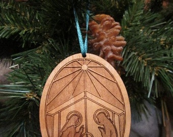 May include: A wooden Christmas ornament with a light brown finish. The ornament is shaped like an oval and features a laser-cut design of the nativity scene. The design includes Mary, Joseph, and the baby Jesus in a manger. The ornament is hanging from a blue ribbon.