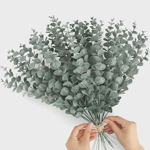 May include: A large bouquet of artificial eucalyptus stems featuring numerous round, dusty green leaves arranged on thin, flexible branches. The stems are gathered together at the base and secured with a piece of natural brown twine.